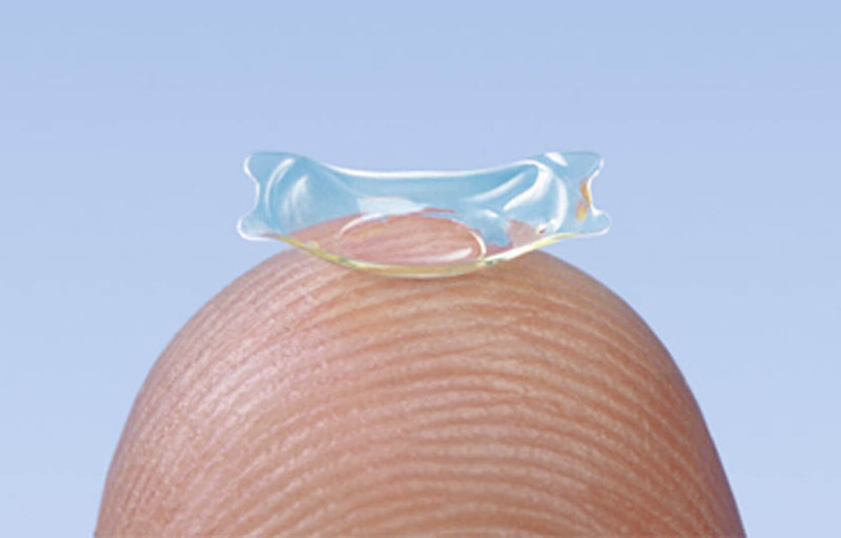 Nahaufnahme einer implantierbaren Kontaktlinse auf einer Fingerspitze vor blauem Hintergrund.