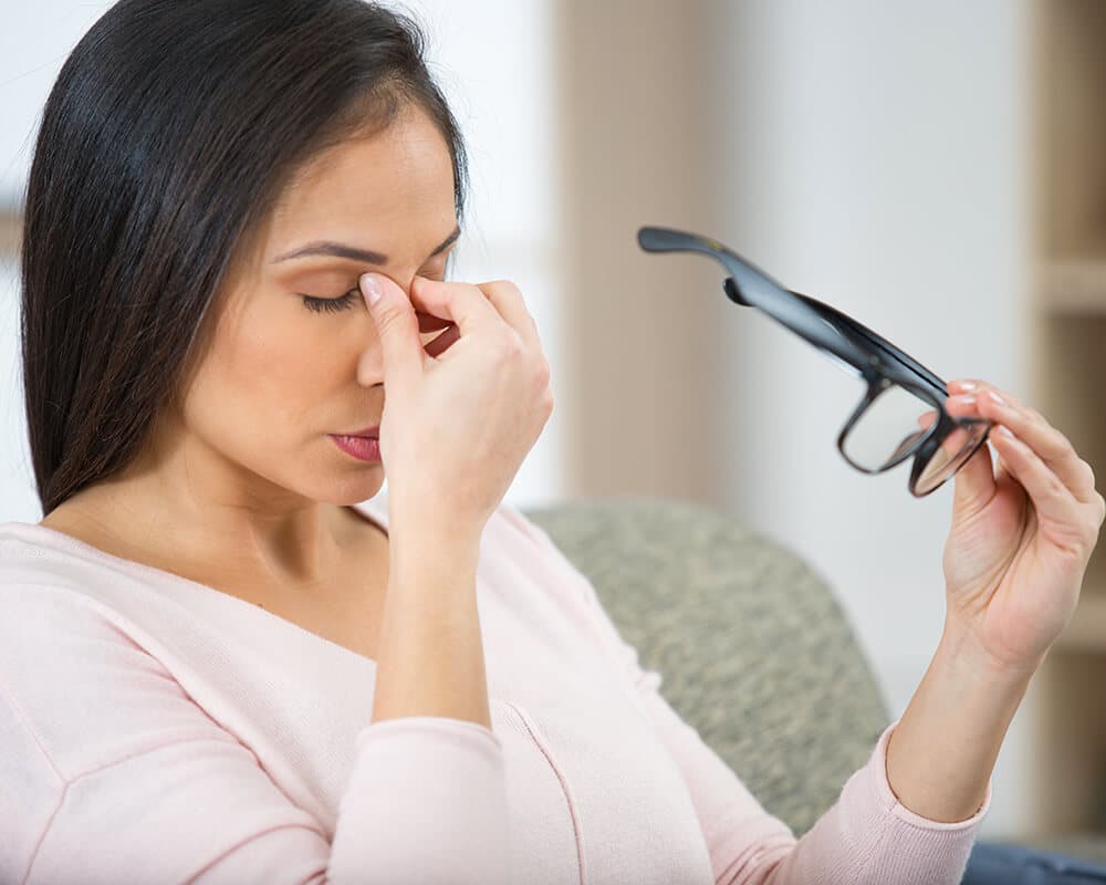 Frau mit Brille reibt sich erschöpft die Augen auf einem Sofa.