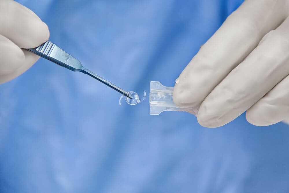Nahaufnahme einer Hand in OP-Handschuhen, die mit einer Pinzette eine Augenlinse hält, vor blauem Hintergrund.