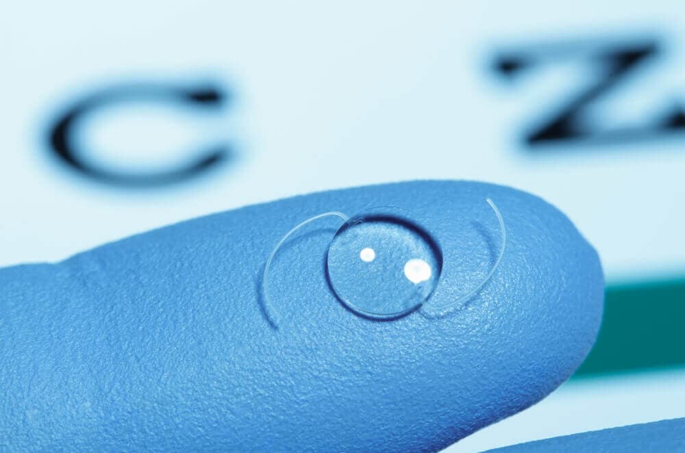 Nahaufnahme einer Implantierbaren Kontaktlinse auf einem blauen behandschuhten Finger, unscharfer Sehtest im Hintergrund.